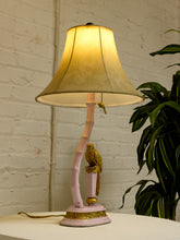Load image into Gallery viewer, A pink table lamp with a curved bamboo-style stem, a gold bird figurine, and a beige fabric bell-shaped shade, shown lit on a white surface against a white brick wall with a green plant to the side.