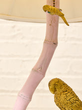 Load image into Gallery viewer, A decorative lamp featuring a pale pink faux-bamboo pole with a small yellow-green parrot figurine perched near the top and another parrot figurine at the lower right, set against a white brick wall and cream lampshade.