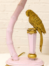 Load image into Gallery viewer, A decorative lamp featuring a gold-colored parrot perched on a pale pink cylindrical post, with a matching pale pink curved bamboo-like arm rising from a round pink base against a white brick wall background.