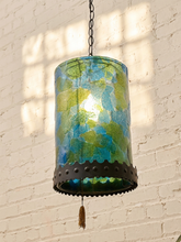 Load image into Gallery viewer, A hanging pendant lamp with a cylindrical glass shade featuring a mottled blue and green pattern, a dark metal scalloped trim, and a tassel, shown against a white brick wall with window light shadows.