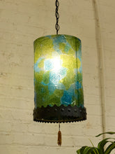 Load image into Gallery viewer, A hanging pendant lamp with a cylindrical glass shade featuring a marbled pattern of green, blue, and yellow tones, illuminated from within. The shade is mounted on a black metal bracket with a decorative tassel hanging below, set against a white brick wall background.