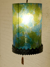 Load image into Gallery viewer, A cylindrical hanging lampshade with a textured, marbled pattern in green, blue, and yellow, illuminated from within. It features a black scalloped metal band with rivets near the bottom and a brown tassel.