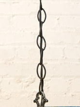 Load image into Gallery viewer, A close-up of a dark metal hanging chain with interlocking oval links and decorative bracket hardware at the bottom, set against a light off-white brick wall background.