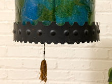 Load image into Gallery viewer, Close-up of a hanging lamp shade with a blue and green marbled top section, a black scalloped metal band with round studs, and a brown tassel pull chain, set against a white brick wall.