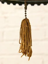 Load image into Gallery viewer, A close-up of a hanging braided tassel ornament in a warm golden-brown color, suspended against a softly blurred white brick wall background.