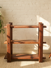 Load image into Gallery viewer, A rustic three-tier wooden shelf with a warm medium-brown finish and brass-colored rivets, set against a white brick wall with soft sunlight and shadows.