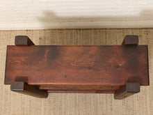 Load image into Gallery viewer, A rustic dark brown wooden bench with thick blocky legs, photographed from directly above on a woven jute rug against a white brick wall.