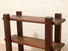 Load image into Gallery viewer, A dark brown wooden three-tier shelf with a rustic industrial design, featuring thick square posts and metal rivets, set against a white brick wall background.