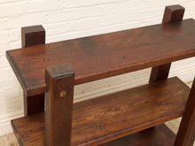 Load image into Gallery viewer, A dark brown wooden three-tier shelf with a rustic industrial design, featuring thick rectangular legs and a visible brass nail on one post, set against a white brick wall.