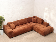 Load image into Gallery viewer, A modular L-shaped sectional sofa upholstered in a textured rust-brown fabric, styled in a minimalist room with white brick walls and a light beige rug.