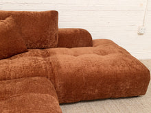 Load image into Gallery viewer, A close-up of a rust-brown corduroy sectional sofa with matching cushions, set against a white brick wall and light carpet.