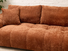 Load image into Gallery viewer, A close-up of a rust-brown corduroy sofa with matching throw pillows, set against a white brick wall background.