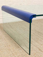 Load image into Gallery viewer, A close-up of a modern glass table corner with a thick blue top rail and dark green metal legs on a beige carpeted floor.