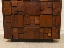 Load image into Gallery viewer, A close-up of a mid-century modern wooden cabinet or dresser featuring a geometric, block-like pattern of dark brown wood panels and a black metal base, set against a white brick wall and light rug.
