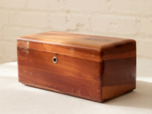 Load image into Gallery viewer, A rectangular wooden storage box with a hinged lid, featuring a warm reddish-brown wood grain finish and a small round metal lock on the front, set against a light brick wall background.