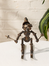 Load image into Gallery viewer, A handmade metal robot figurine crafted from rusty brown screws, nuts, and bolts, holding a small metal key in each hand, standing on a white textured surface against a white brick wall background.