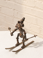 Load image into Gallery viewer, A rustic metal sculpture of a skiing figure made from repurposed gears, springs, and bolts, holding ski poles on a white textured surface against a white brick wall background.