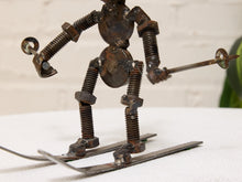 Load image into Gallery viewer, A rustic metal sculpture of a skier made from repurposed bolts, nuts, and screws, holding ski poles and standing on metal skis against a white brick wall background.