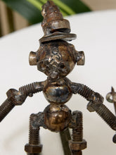 Load image into Gallery viewer, A handmade metal sculpture of a whimsical robot or gnome figure constructed from rusty bolts, nuts, and threaded rods, photographed close-up against a white surface with a blurred green background.