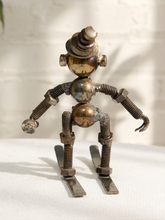 Load image into Gallery viewer, A handmade metal robot figurine made from repurposed industrial parts, including screws, nuts, washers, and a brass-toned sphere, posed standing on metal plates against a white textured background.