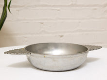 Load image into Gallery viewer, A vintage silver metal bowl with decorative leaf-shaped handles, resting on a white textured surface against a white brick wall background.