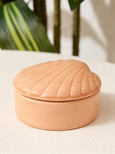 Load image into Gallery viewer, A small round lidded box with a shell-shaped lid, made of terracotta-colored ceramic, sitting on a textured white fabric surface with a blurred green plant in the background.