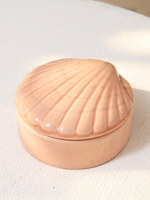 Load image into Gallery viewer, A small round lidded container shaped like a scallop shell, in a glossy peach-pink color, resting on a textured off-white fabric surface.