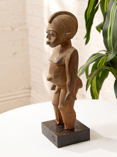 Load image into Gallery viewer, A small hand-carved wooden sculpture of a stylized human figure with a dark rectangular base, displayed on a white table with a white brick wall and a green plant in the background.