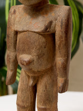 Load image into Gallery viewer, A close-up of a hand-carved wooden figurine with a textured, weathered brown finish, set against a soft-focus background of green leaves and a neutral wall.