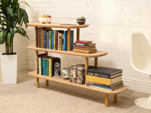 Load image into Gallery viewer, A three-tier wooden bookshelf with a light natural oak finish, styled with books, a plant, and a chair in a bright room.
