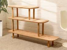 Load image into Gallery viewer, A three-tier wooden shelving unit with a natural light oak finish, styled in a room setting against a white brick wall with a plant and chair partially visible.