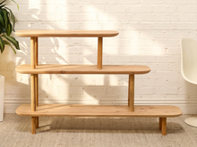 Load image into Gallery viewer, A three-tier wooden step shelf with a natural light oak finish, set against a white brick wall with soft leaf shadows.