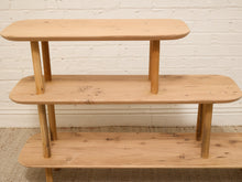 Load image into Gallery viewer, A three-tier wooden shelf unit made from light natural pine wood, set against a white painted brick wall and a beige carpet.