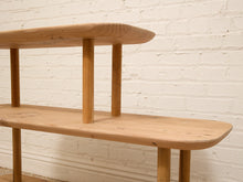 Load image into Gallery viewer, A close-up of a light natural wood bench or shelf with rounded ends and cylindrical legs, set against a white painted brick wall.