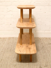 Load image into Gallery viewer, A three-tier wooden display shelf with a natural light oak finish, standing on a textured beige rug against a white painted brick wall.