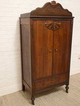 Load image into Gallery viewer, A tall vintage wooden armoire cabinet with ornate carved details on the doors and top, standing against a white painted brick wall on a beige carpet.