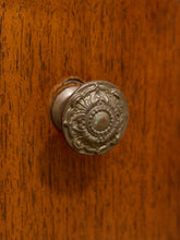 Load image into Gallery viewer, An ornate round metal cabinet knob with a floral relief design, mounted on a warm brown wooden door.