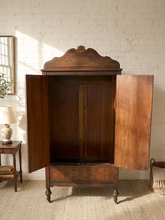 Load image into Gallery viewer, An antique dark wood armoire cabinet with its doors open, set against a white brick wall in a styled room with a side table and lamp.