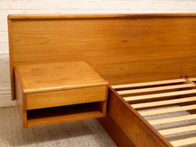 Load image into Gallery viewer, A mid-century modern wooden bed frame with a matching nightstand and headboard in a warm honey oak finish, shown against a white brick wall and carpet.