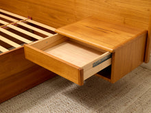 Load image into Gallery viewer, Close-up of a wooden bed frame drawer with the front panel pulled open, showing the light wood interior and metal drawer pull hardware, set against a neutral carpeted floor.