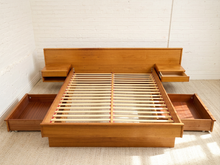 Load image into Gallery viewer, A mid-century modern wooden platform bed frame with an open headboard, slatted support base, and four pull-out storage drawers, shown in a bright room with white brick walls and light-colored carpet.