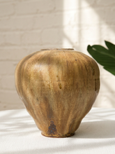 Load image into Gallery viewer, A rustic ceramic vase with a textured, organic shape in earthy brown and gold tones, sitting on a white fabric surface against a white brick wall with soft window shadows.