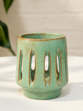Load image into Gallery viewer, A ceramic candle holder with a light green glaze and a rustic brown distressed finish, featuring vertical oval cutouts, set on a white fabric surface with a blurred background.