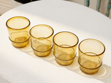Load image into Gallery viewer, Four amber-tinted glass tumblers stacked in a row on a white textured tablecloth, with natural sunlight casting soft shadows.