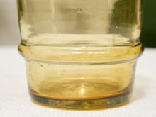 Load image into Gallery viewer, A close-up of a clear glass jar or container with a thick, rounded base, showing a warm amber-yellow tint and small air bubbles trapped inside the transparent material.