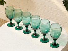 Load image into Gallery viewer, Six translucent teal-green wine glasses arranged in a row on a white round table against a white brick wall background.