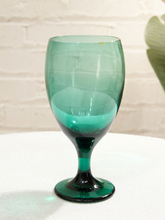 Load image into Gallery viewer, A translucent green glass goblet with a dark green base, standing on a white textured surface against a white brick wall background.