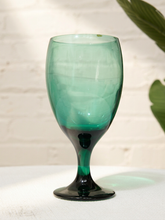 Load image into Gallery viewer, A translucent teal green goblet glass with a dark green base, set on a white textured surface against a white brick wall with soft leaf shadows.