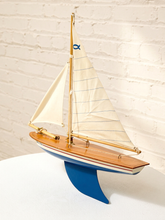 Load image into Gallery viewer, A wooden model sailboat with cream-colored sails, a blue hull stripe, and a blue support stand, displayed against a white brick wall background.