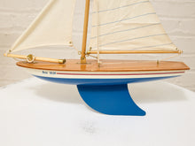 Load image into Gallery viewer, A wooden model sailboat with cream-colored sails, a polished wood hull, and a blue support stand, photographed against a white brick wall background.